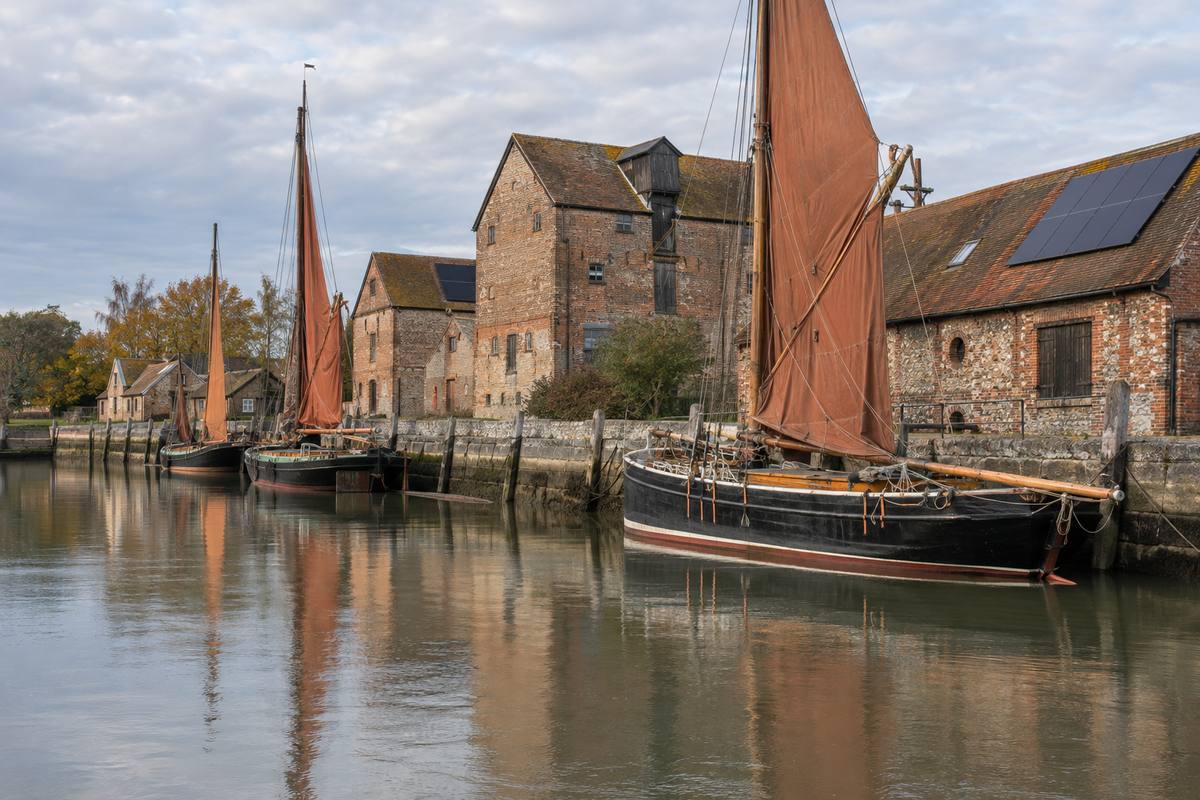 Thames sailing barges with ochre sails moored at a tidal quay in front of old brick and flint warehouses, with solar panels on two roofs, under an autumn sky